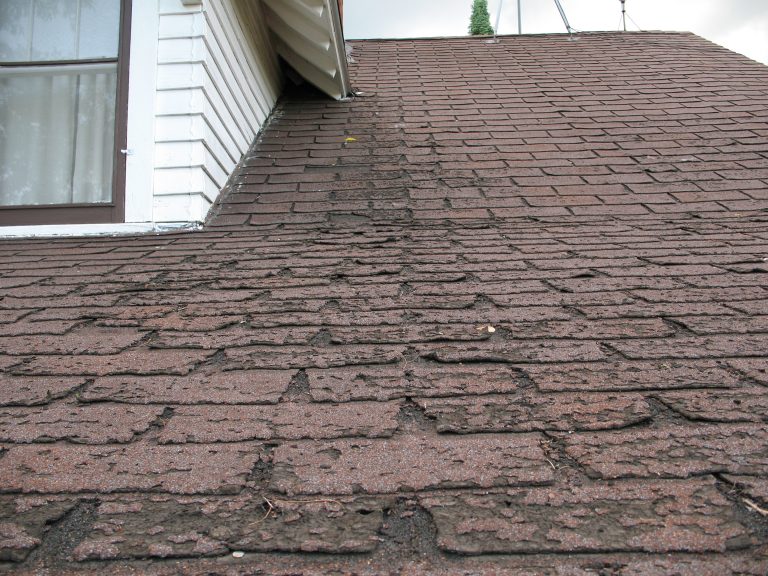 Close-up of a roof showing widespread shingle damage. The granules have worn away and the asphalt layer underneath is exposed, indicating the need for repair or replacement